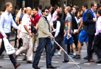 Un hombre ciego con un baston blanco está cruzando la calle junto a otros peatones